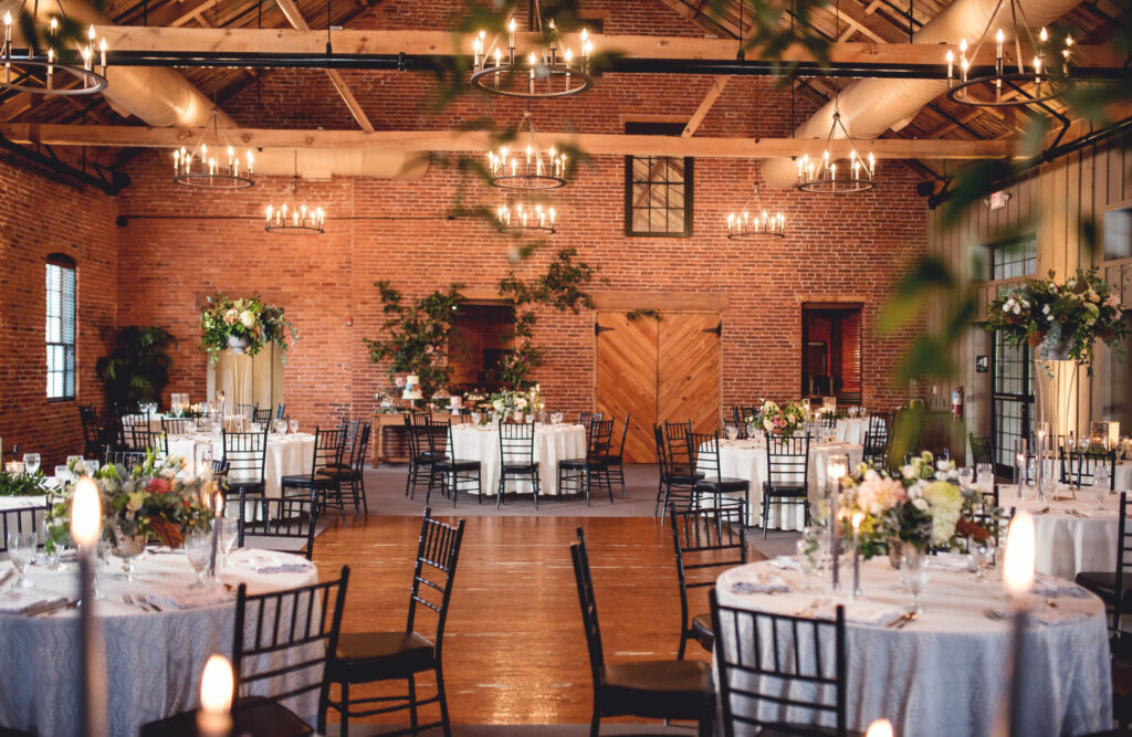 Elegantly decorated event hall with round tables, white tablecloths, floral centerpieces, and black chairs. Brick walls and chandeliers create a warm ambiance.