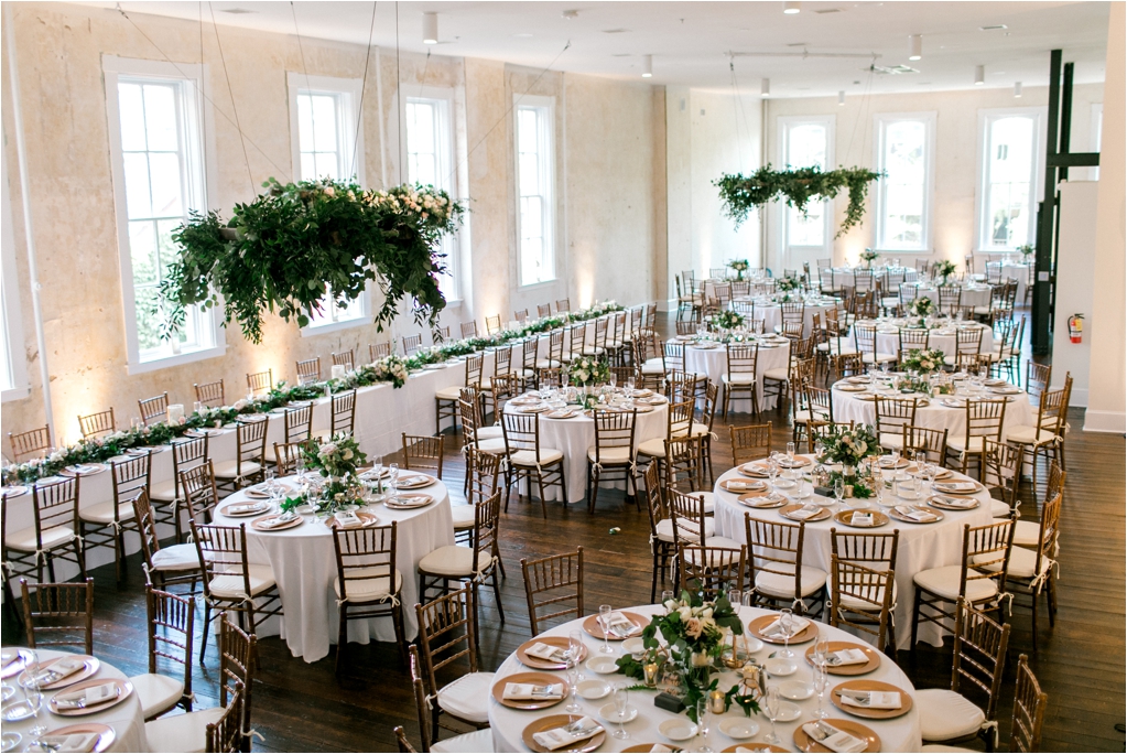 Elegant wedding reception setup with round tables, wooden chairs, and hanging greenery. Tables are set with white tablecloths, gold-rimmed plates, and floral centerpieces. Large windows in the background.