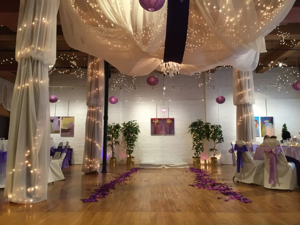 A wedding venue with wooden flooring, draped white fabric and string lights from the ceiling, purple decorations, and dining tables. A path lined with purple petals leads to a brick wall.