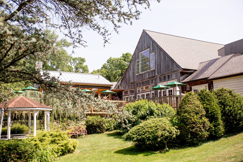 Large rustic building with wooden exterior, surrounded by lush green trees and shrubs. A gazebo and outdoor seating area with umbrellas are visible nearby under a clear sky.