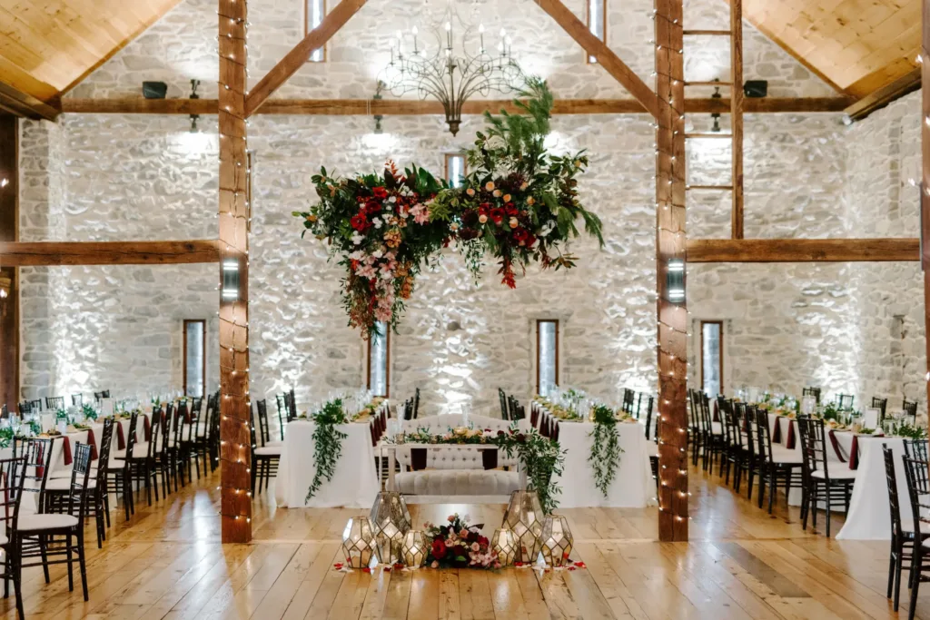 Barn wedding venue with wooden beams and stone walls, featuring a floral chandelier centerpiece, long tables with greenery, wooden chairs, and lanterns on the floor.