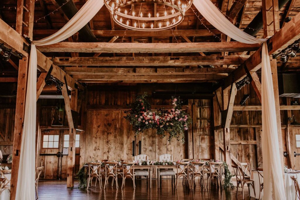 Rustic wooden barn interior set for an event with draped fabric, a chandelier, floral centerpiece, and long table surrounded by wooden chairs.