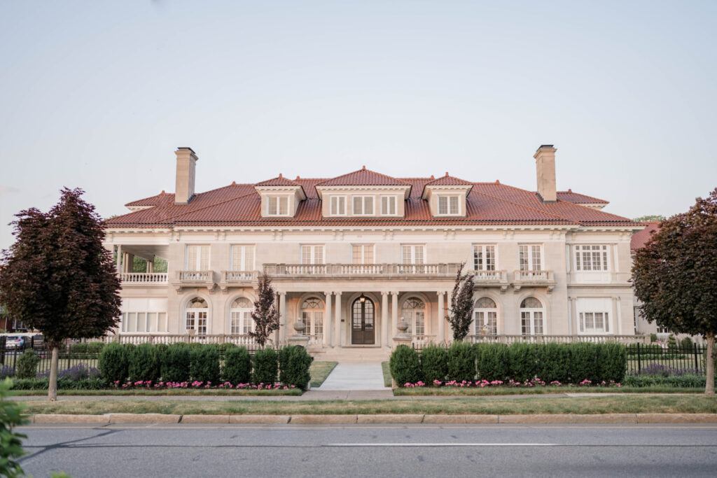 A large, symmetrical mansion with a red-tiled roof, multiple chimneys, arched windows, and a landscaped front yard bordered by bushes and flowers—perfect for hosting an elegant wedding.