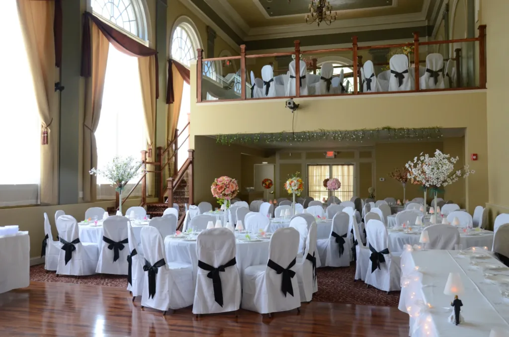 A banquet hall decorated for a wedding event with round tables covered in white cloths, black chair sashes, floral centerpieces, large windows, and an upper balcony area.