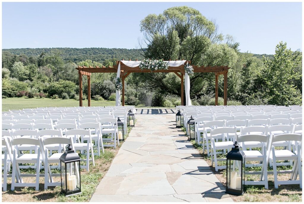 Outdoor wedding ceremony setup with rows of white chairs facing a wooden pergola decorated with white fabric and greenery, surrounded by nature. Large lanterns line the aisle, creating a romantic atmosphere for the perfect wedding celebration.