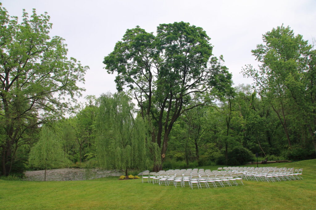 Rows of white folding chairs are set up outdoors on grass beside a pond, surrounded by green trees under a cloudy sky, ready for a picturesque wedding ceremony.