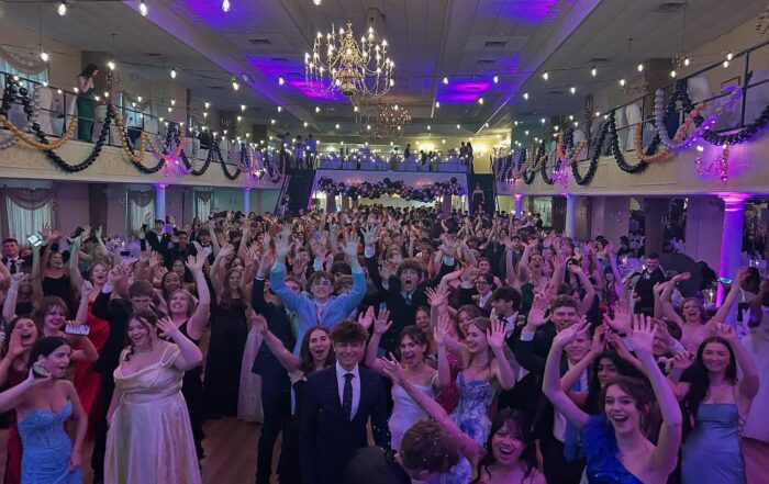 A large group of people in formal attire stand and cheer with their hands raised at a decorated indoor event, with a DJ keeping the energy high—perfect for a memorable prom, PA wedding, or special occasion in Gettysburg PA.