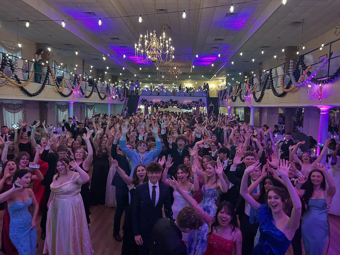 A large group of people in formal attire stand and cheer with their hands raised at a decorated indoor event, with a DJ keeping the energy high—perfect for a memorable prom, PA wedding, or special occasion in Gettysburg PA.