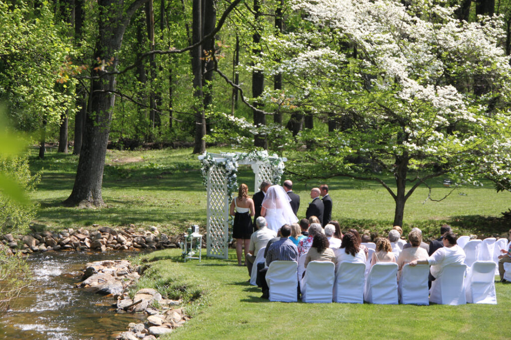 Outdoor wedding ceremony by a creek at one of the charming Gettysburg wedding venues, with the bride and groom standing under an arbor and guests seated on white chairs on a sunny day.