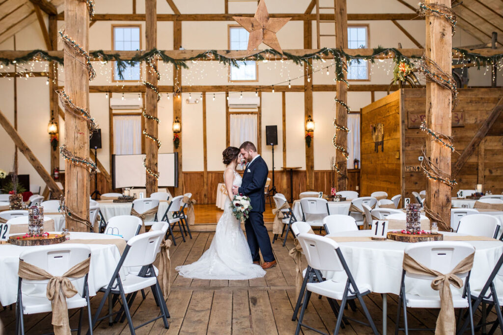 A bride and groom stand together in the center of a rustic, decorated barn venue—one of the charming Gettysburg wedding venues—surrounded by round tables with white tablecloths and brown accents.