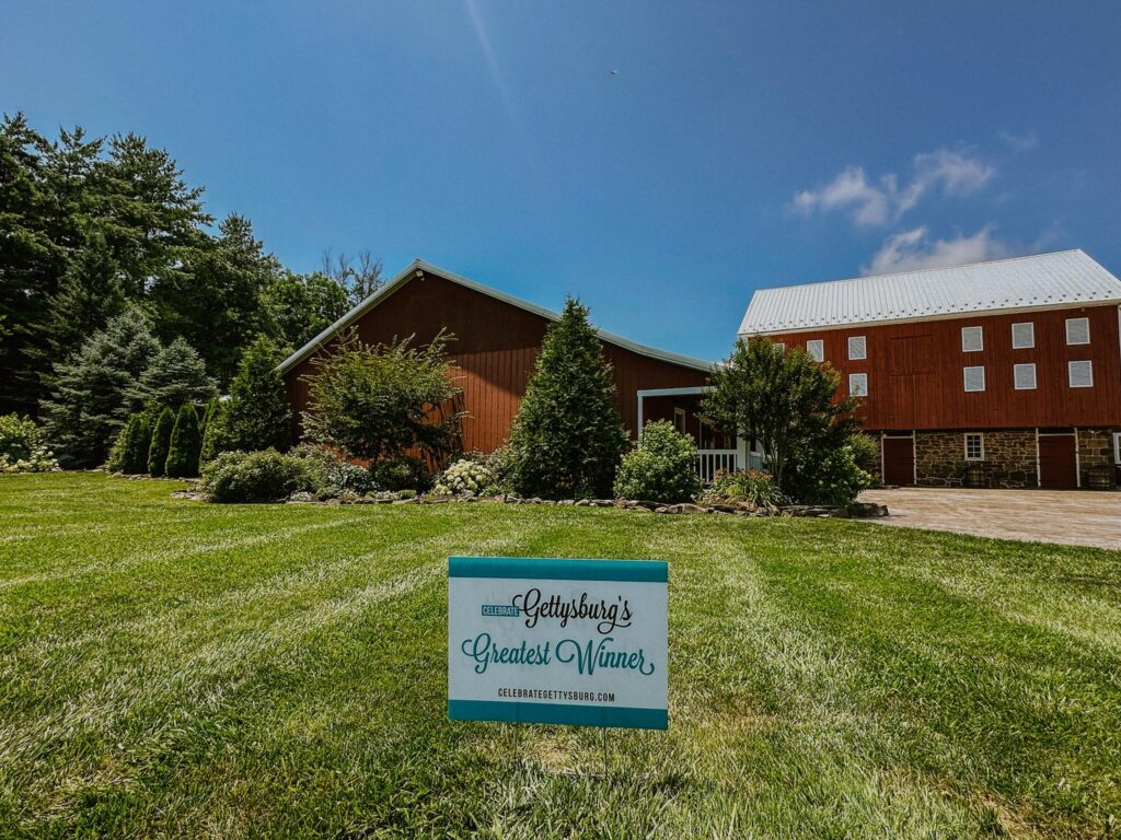 A large red barn sits on a well-manicured lawn with a sign in the grass reading “Gettysburg’s Greatest Winner,” making it one of the most charming Gettysburg wedding venues, all beneath a stunning blue sky.