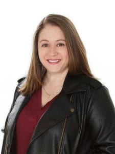 A woman with straight, brown hair wearing a black leather jacket and a maroon top smiles at the camera against a plain white background, exuding the confident vibe of an event DJ ready to set the mood.