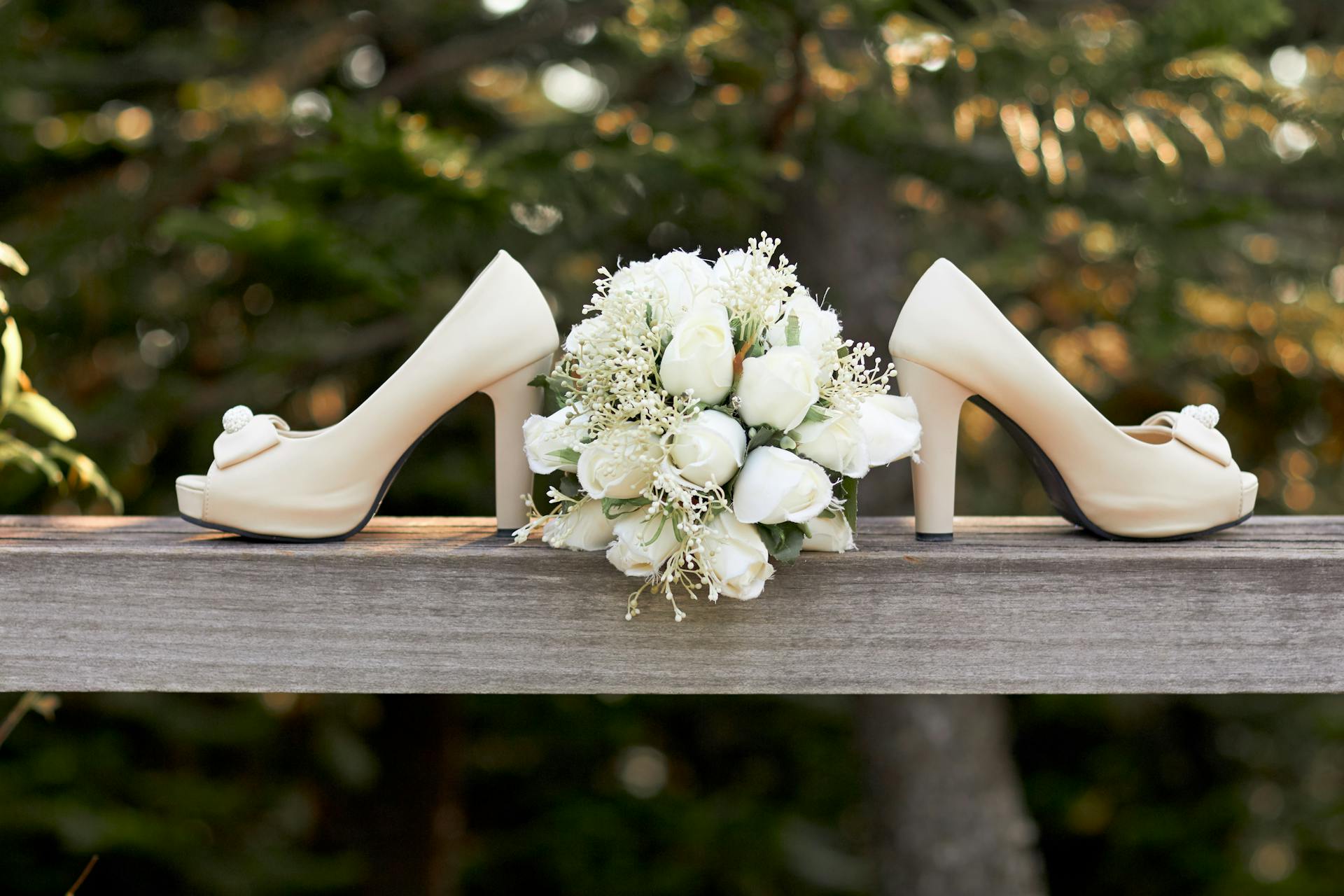 A pair of cream-colored high-heeled shoes and a white rose bouquet are displayed on a wooden rail outdoors, surrounded by greenery—perfect inspiration for celebrations at the top wedding venues in Lancaster PA.