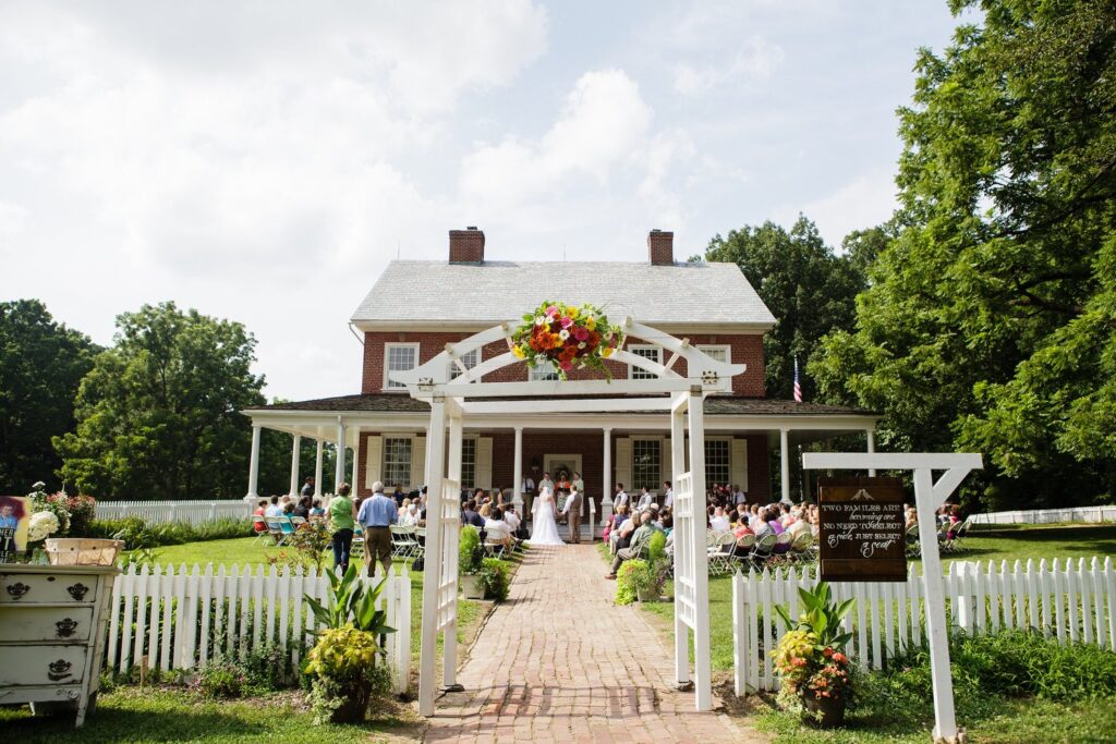 An outdoor wedding ceremony unfolds at one of the top wedding venues in Lancaster PA, with guests seated beside a brick walkway and a flower-adorned archway set before a charming historic house.