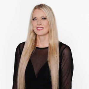 A woman with long blonde hair wearing a black sheer-sleeved top stands against a plain white background, smiling warmly as if about to share something about herself.