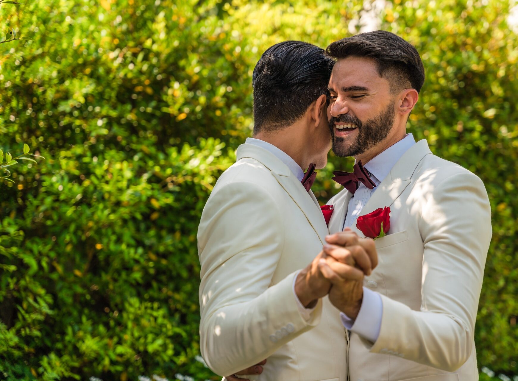 Two men in matching white suits with red bow ties and pocket squares dance together outside, surrounded by greenery, as an LGBTQ wedding DJ sets the joyful mood.
